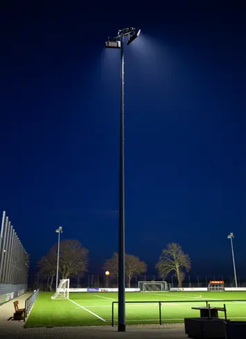 Ein beleuchteter Flutlichtmast auf einem Fußballplatz bei Nacht. Der Rasen ist grün und gut ausgeleuchtet, während der Himmel dunkelblau ist. Im Hintergrund stehen Bäume und ein Tor, teilweise von der Beleuchtung angestrahlt. Links befindet sich ein Zaun.