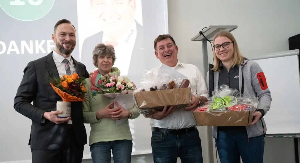 Vier Personen lächeln und halten Geschenke in Form von Blumensträußen und Geschenkkörben vor einem Präsentationshintergrund mit 'Danke'-Text.