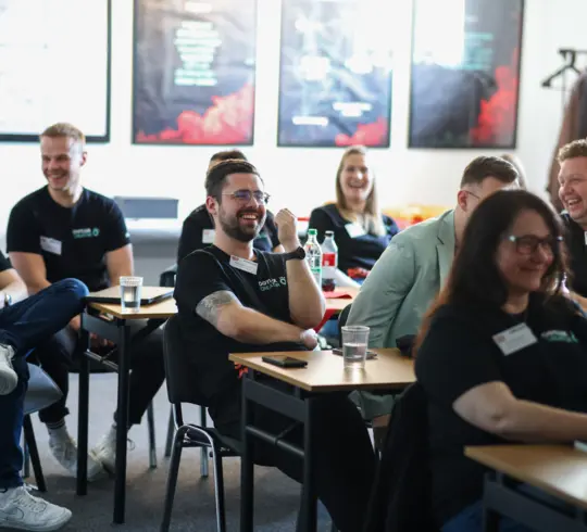 Eine Gruppe von Personen in einem modernen Schulungsraum lacht und wirkt entspannt, während sie an einem Workshop oder einer Teamveranstaltung teilnehmen. Sie sitzen an kleinen Tischen mit Getränken und Arbeitsmaterialien, die Atmosphäre ist locker und konzentriert.