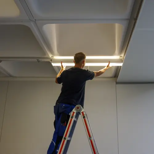 Ein Arbeiter auf einer Leiter installiert eine QUICK-FIXdc Deckenlampe in einem Raum mit moderner Rasterdecke.