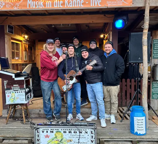 Eine Gruppe von Männern steht zusammen auf einer kleinen Bühne vor einem Banner mit der Aufschrift „Musik in die Kanne – live“. Einer hält eine Gitarre, während die anderen lächeln. Im Vordergrund ist ein Aufsteller mit der Aufschrift „Live Music Entertainment Tim Brown“.