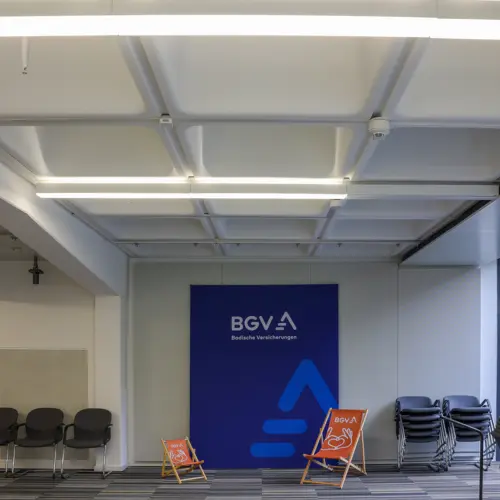 Ein Raum mit Rasterdecke, hellen Lampen, zwei orange "BGV"-Liegestühlen und einem blauen Banner der "BGV Badische Versicherungen".