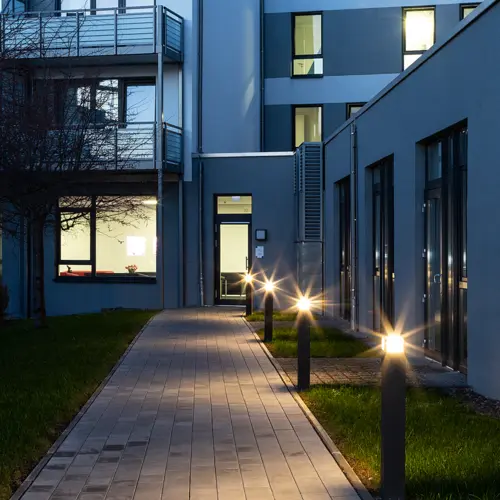 Abendaufnahme eines gepflasterten Gehwegs, flankiert von beleuchteten WAY-Leuchten, vor einem modernen Gebäude mit großen Fenstern.