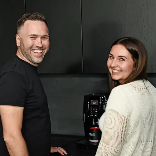 Ein Mann und eine Frau lachen herzlich, während sie sich in einer modernen Küche an einer Kaffeemaschine befinden.