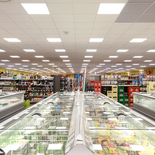 Ein heller Supermarkt mit Reihen von Kühltruhen in der Mitte, flankiert von gut sortierten Regalen mit Getränken und Lebensmitteln. Die Decke ist mit vielen rechteckigen LED-Panels beleuchtet.