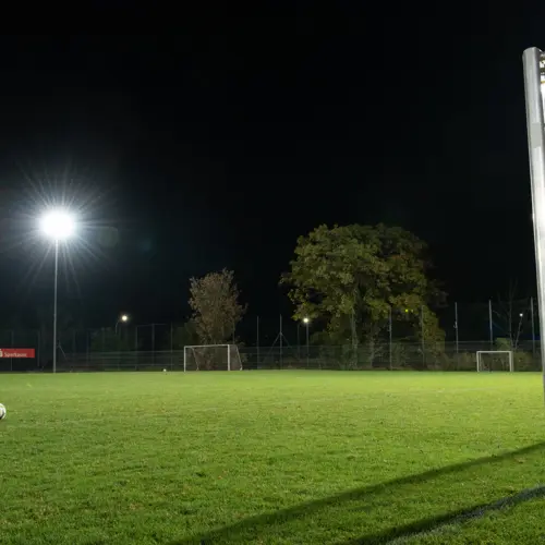 Ein beleuchteter Fußballplatz bei Nacht mit einem Flutlichtmast links, das den Rasen erhellt. Im Hintergrund sind Bäume und zwei Tore sichtbar.