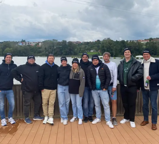 Eine Gruppe von zehn Personen steht auf einer Holzplattform vor einem See. Die meisten tragen schwarze Mützen mit dem Schriftzug „Dotlux“. Im Hintergrund ist eine bewaldete Uferlandschaft mit einigen Häusern zu sehen, der Himmel ist bewölkt.