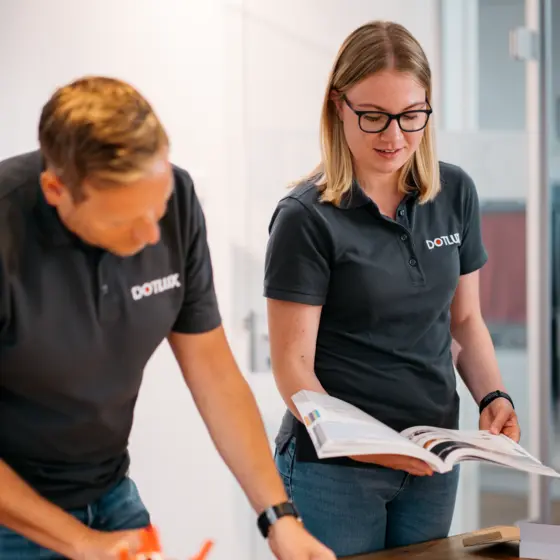 Eine Frau im DOTLUX-Outfit betrachtet ein aufgeschlagenes Katalogheft, während ein Kollege neben ihr an einem Tisch arbeitet.