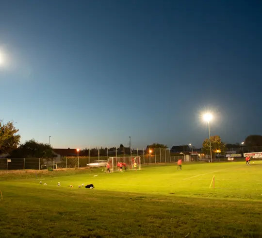 Fußballplatz bei Dämmerung, beleuchtet von Flutlichtanlagen. Spieler trainieren am Tor, während Fußballbälle entlang der Seitenlinie verteilt sind.