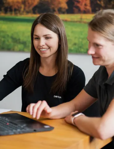 Zwei Personen bei der Zusammenarbeit an einem Laptop, im Hintergrund eine idyllische Landschaft auf einem Bild.