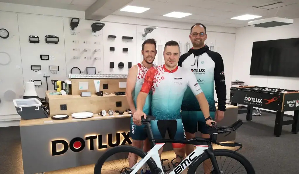 Drei Männer in DOTLUX-Radsportbekleidung stehen mit einem Rennrad in einem Showroom voller Beleuchtungsprodukte, neben einem DOTLUX-gebrandeten Tischkicker.