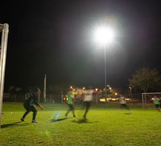 Nachtaufnahme eines Fußballtrainings auf einem hell beleuchteten Spielfeld. Spieler in Bewegung sind leicht unscharf, während die Flutlichtanlage und Umgebung klar zu erkennen sind.