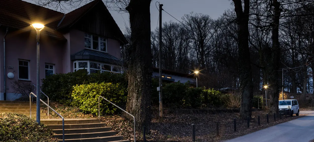 Eine ruhige, beleuchtete Straße in der Dämmerung, gesäumt von Bäumen und mit einem Wohnhaus im Hintergrund.